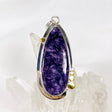 Purple Charoite tear drop pendant with brass detailing in sterling silver sitting on a crystal cluster