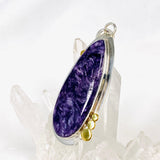 Purple Charoite tear drop pendant with brass detailing  in sterling silver sitting on a crystal cluster