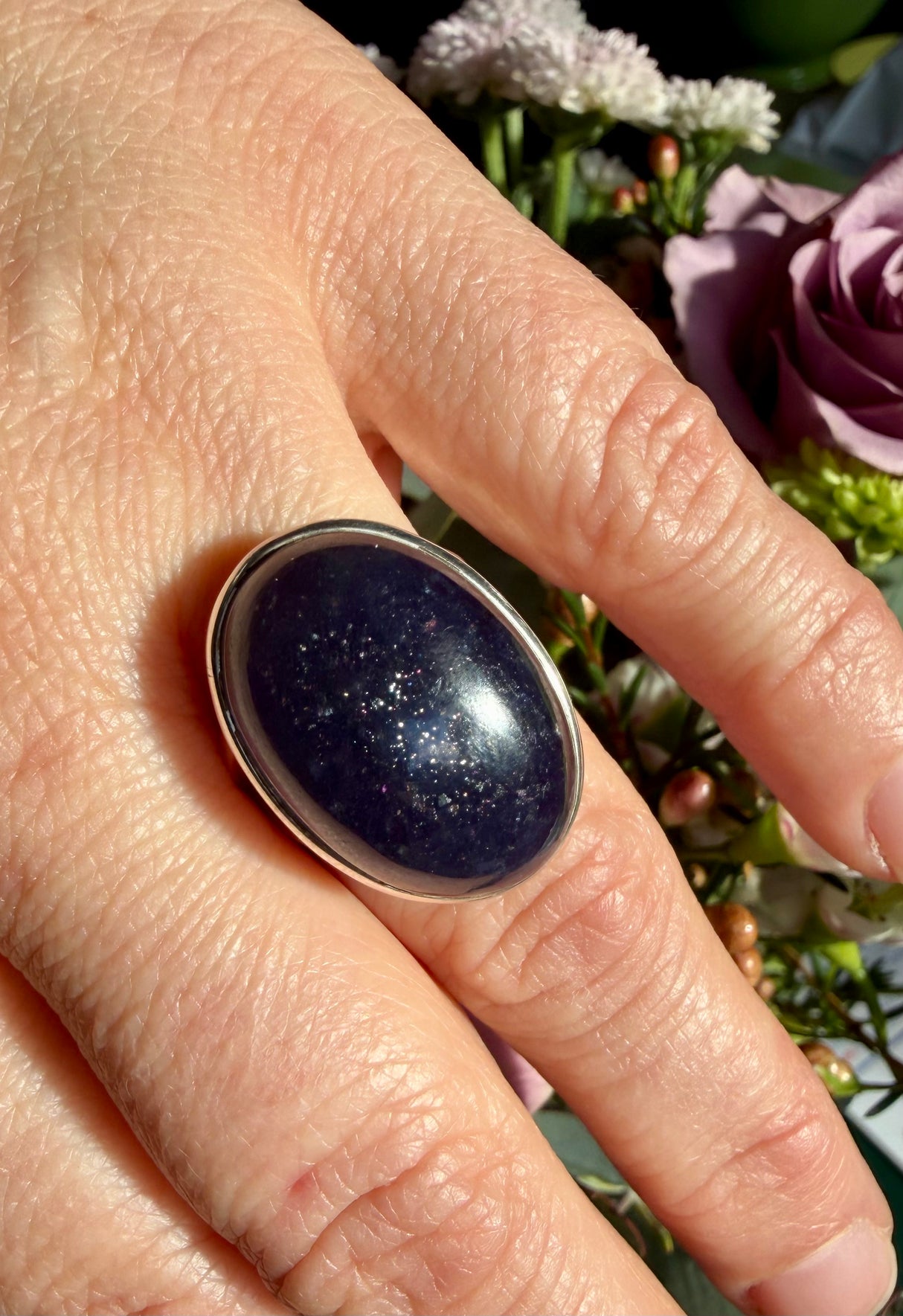 Sunstone in iolite oval cabochon ring worn on the hand against a floral background, showing deep indigo-violet colour with a cluster of fine sparkle catching the light