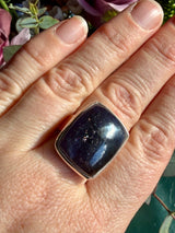 Sunstone in iolite rectangular cabochon ring worn on the hand against a floral background, showing deep indigo-purple colour with a dense cluster of sparkle catching the light