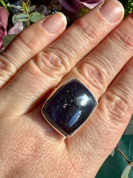 Sunstone in iolite rectangular cabochon ring worn on the hand against a floral background, showing deep indigo-purple colour with a dense cluster of sparkle catching the light