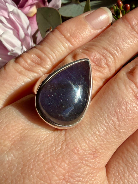 Sunstone in iolite teardrop cabochon ring worn on the hand against a floral background, showing deep indigo-blue-violet colour with scattered sparkle