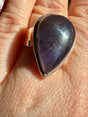 Sunstone in iolite teardrop cabochon ring worn on the hand against a floral background, showing deep indigo-violet tones with scattered sparkle and a bright glint