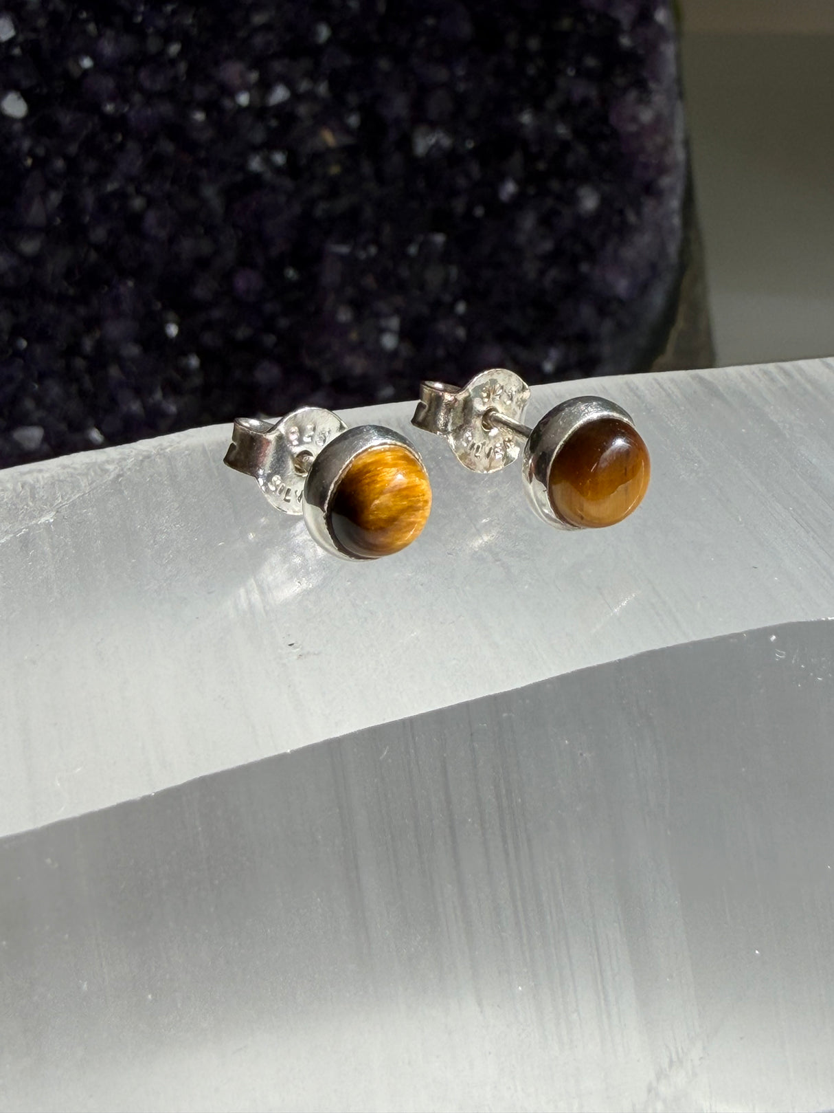A pair of stud earrings with round brown tiger's eye gemstones in silver bezel settings, displayed on a white crystal slab with a dark purple crystal cluster in the background.