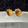 A pair of silver stud earrings with round, polished golden-brown tiger's eye gemstones, displayed on a translucent white crystal block against a dark, sparkly black background.