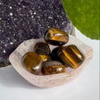 Five polished tiger's eye stones with brown and gold bands, resting in a small translucent pink crystal dish. The dish is placed on a white surface, with a purple crystal cluster on the left and a green leaf on the right.