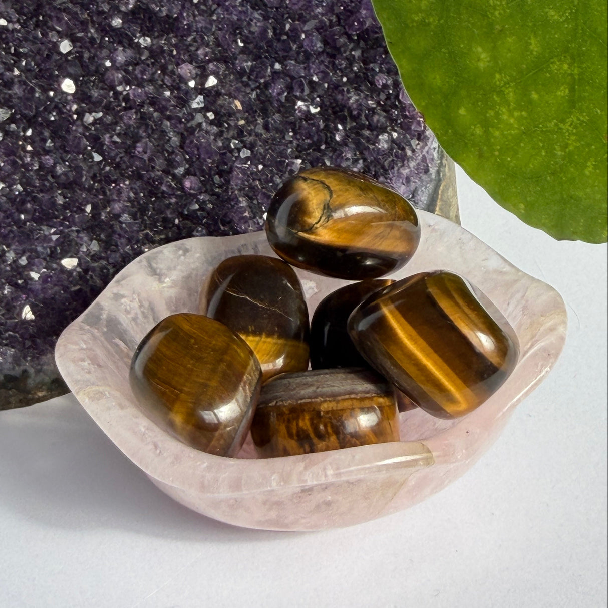 Five polished tiger's eye stones with brown and gold bands, resting in a small translucent pink crystal dish. The dish is placed on a white surface, with a purple crystal cluster on the left and a green leaf on the right.