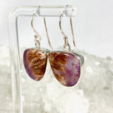 Purple Charoite free form earrings in sterling silver hanging on an earring stand