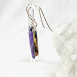 Purple Charoite tear drop earrings in sterling silver hanging on an earring stand