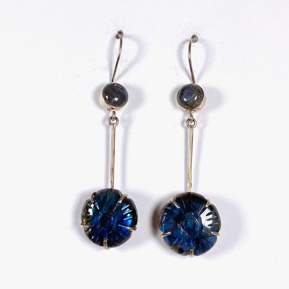 Labradorite and silver earrings with blue flower carving at the bottom with a silver stem going up to a small round labradoite gemstone with a fixed hookl going through a piece of white cardboard