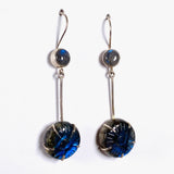 Labradorite and silver earrings with blue flower carving at the bottom with a silver stem going up to a small round labradoite gemstone with a fixed hookl going through a piece of white cardboard